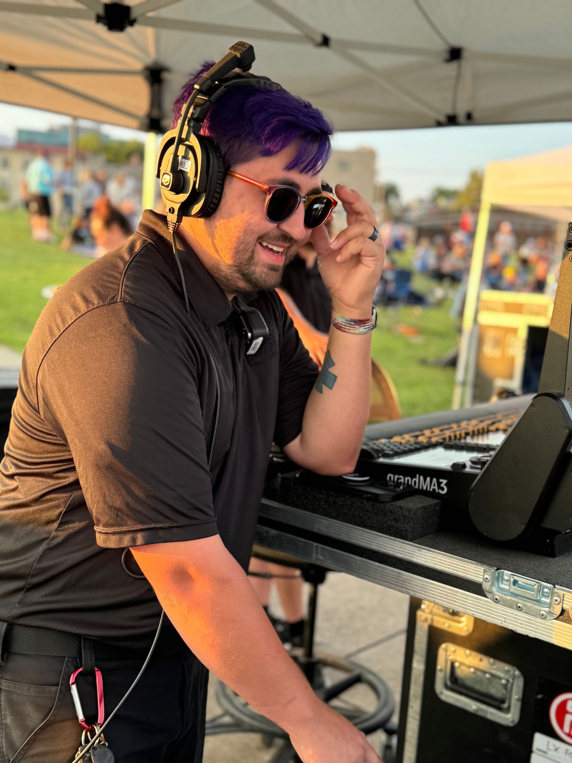 Man with purple hair and sunglasses at an outdoor DJ setup, smiling, wearing headphones, with people in the background.