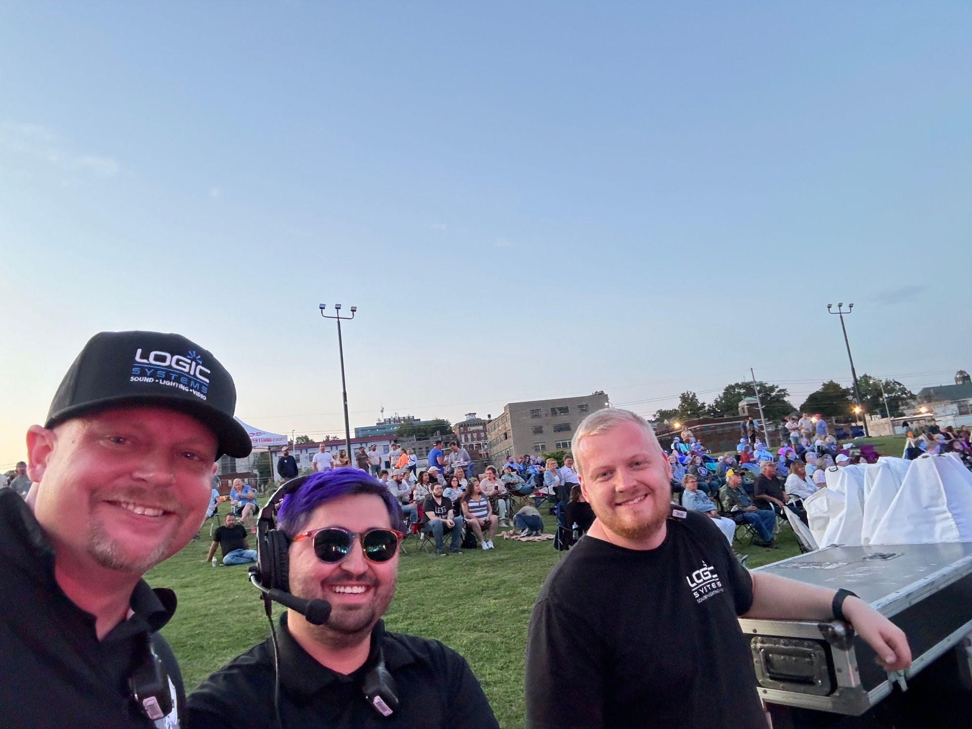 Three men smiling at an outdoor event, crowd in background, at dusk.