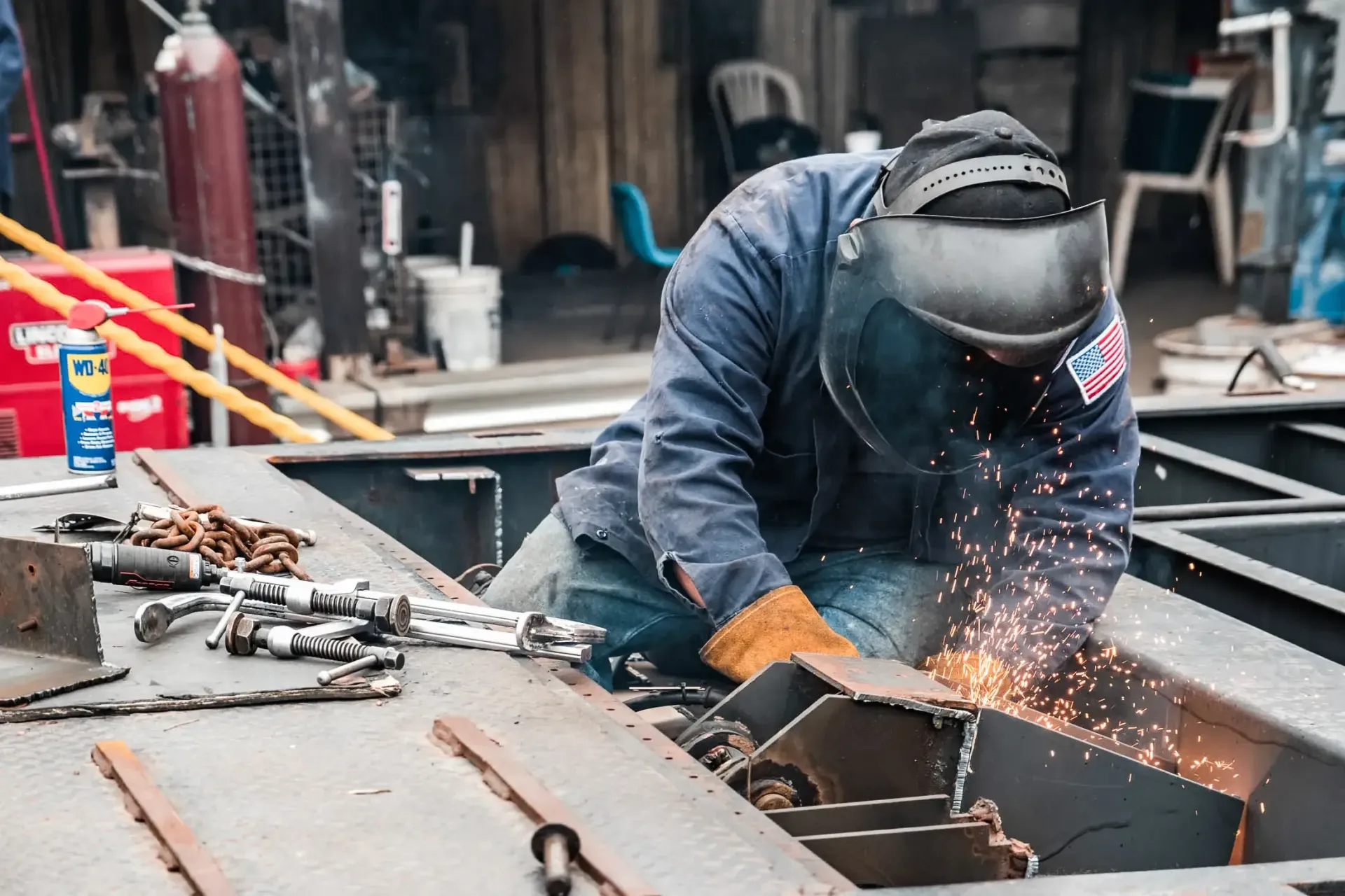 A person wearing a welding helmet and protective gloves welds a metal structure, creating sparks in a workshop.