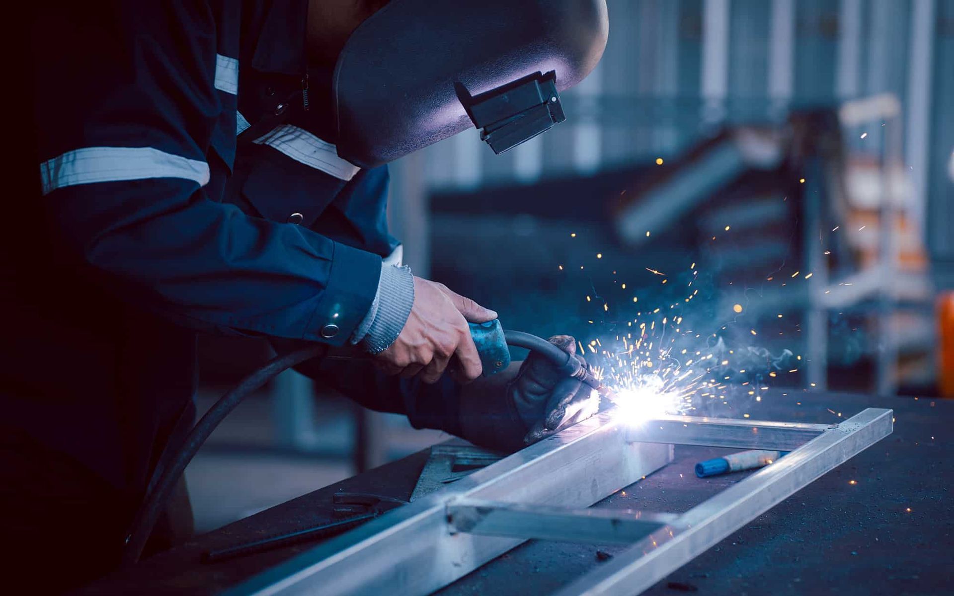 A person wearing a welding helmet and protective gear uses a tool to weld a metal frame, creating sparks in a workshop.