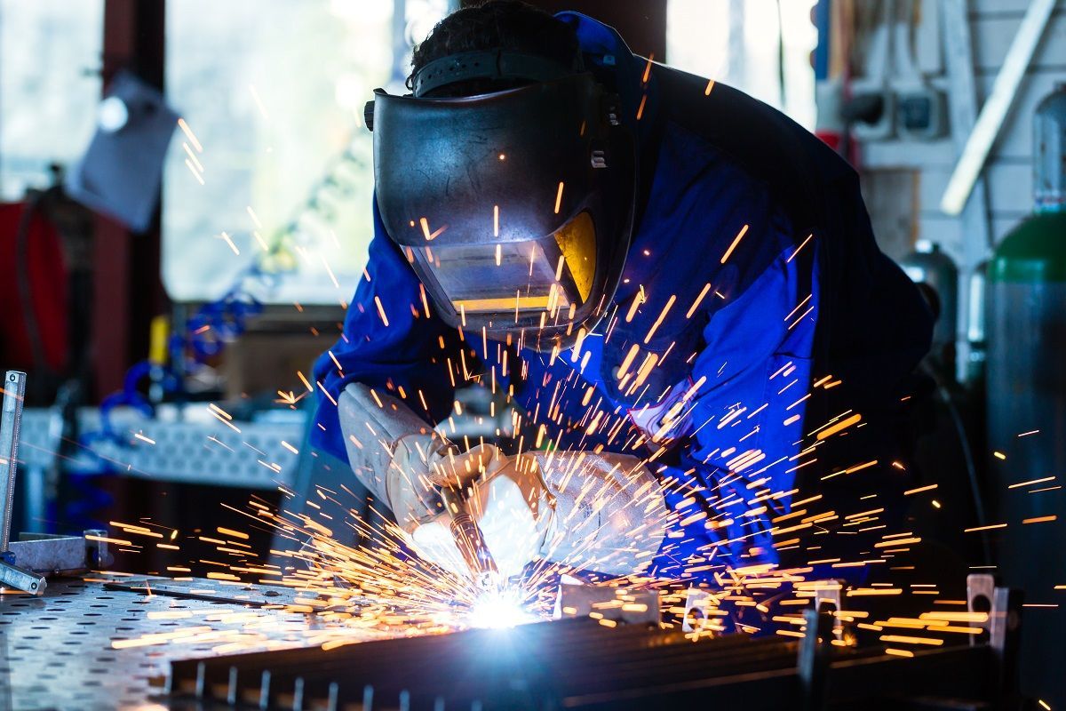A welder wearing protective gear and a mask works on metal, producing a spray of bright orange sparks in a workshop.