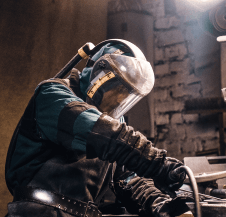 A person wearing a protective face shield, heavy gloves, and apron works with metal tools in a workshop.