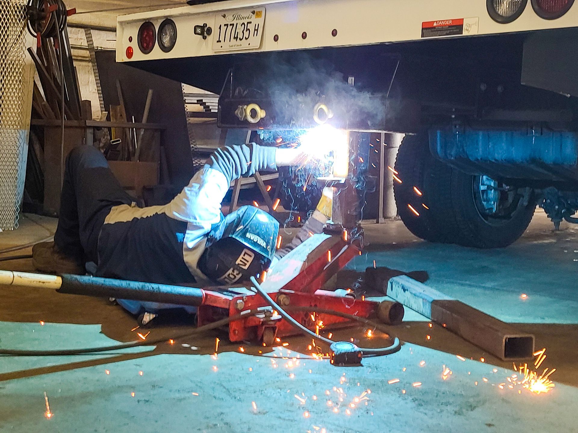 A welder lies on the floor under the back of a truck, performing repairs while sparks fly from the welding torch.