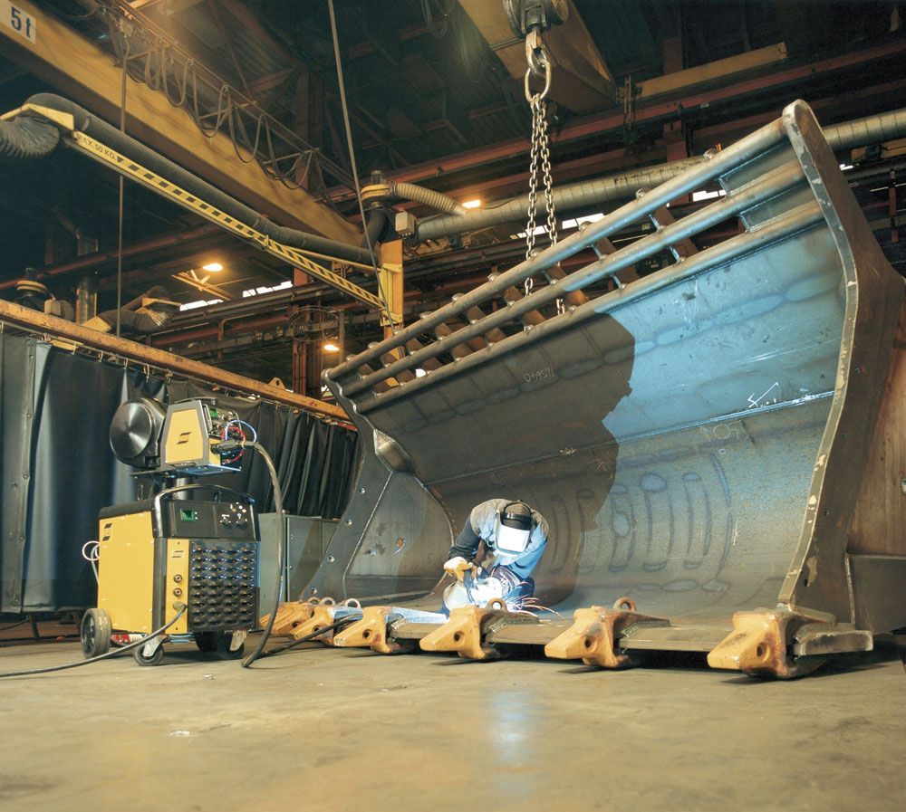 A worker in protective gear welds the interior of a large metal industrial excavator bucket inside a workshop.