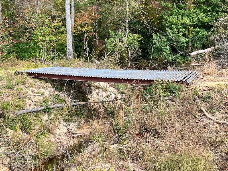 A simple footbridge made of corrugated metal roofing sheets spanning a small, earthen creek bed in a wooded area.