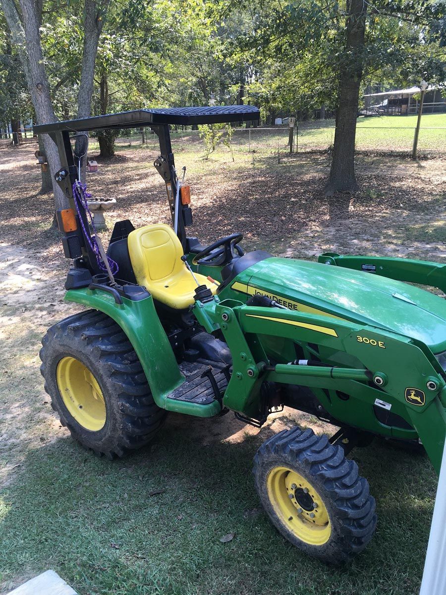 A green John Deere tractor with a yellow seat and front-end loader parked on a dirt path in a wooded, sunny area.