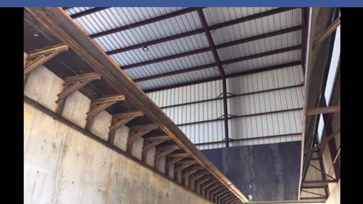 Angled view of an industrial building interior with metal roof panels supported by a concrete wall and steel brackets.