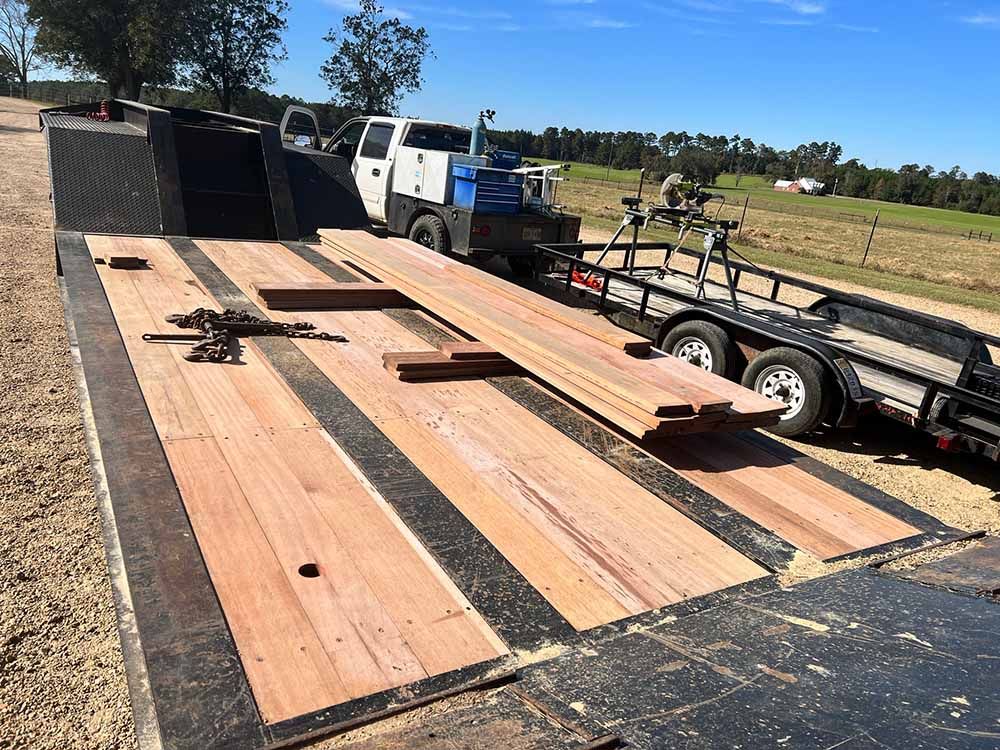 A flatbed trailer parked on a dirt lot, featuring newly installed wooden planks on its deck, with a truck and gear nearby.