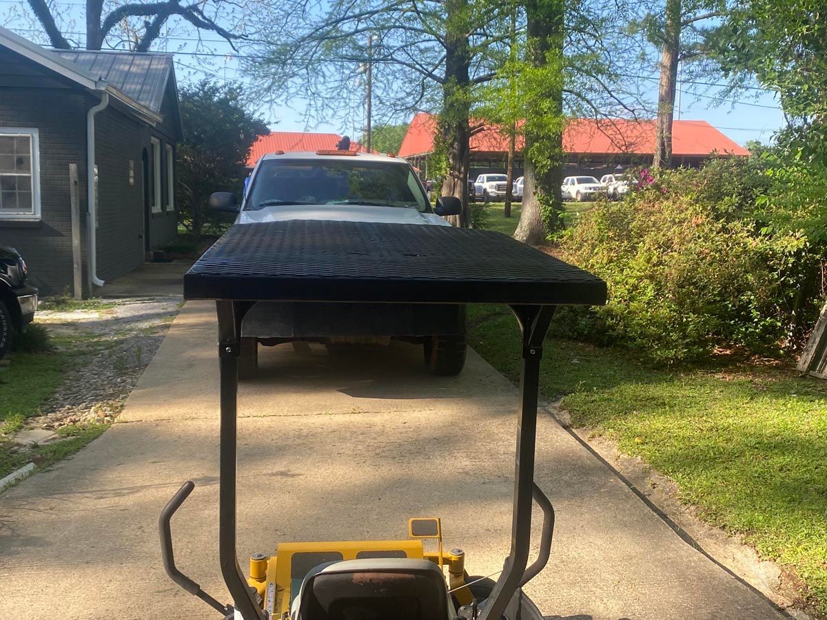 A yellow zero-turn lawn mower with a black canopy in a driveway, with a vehicle parked in the background.