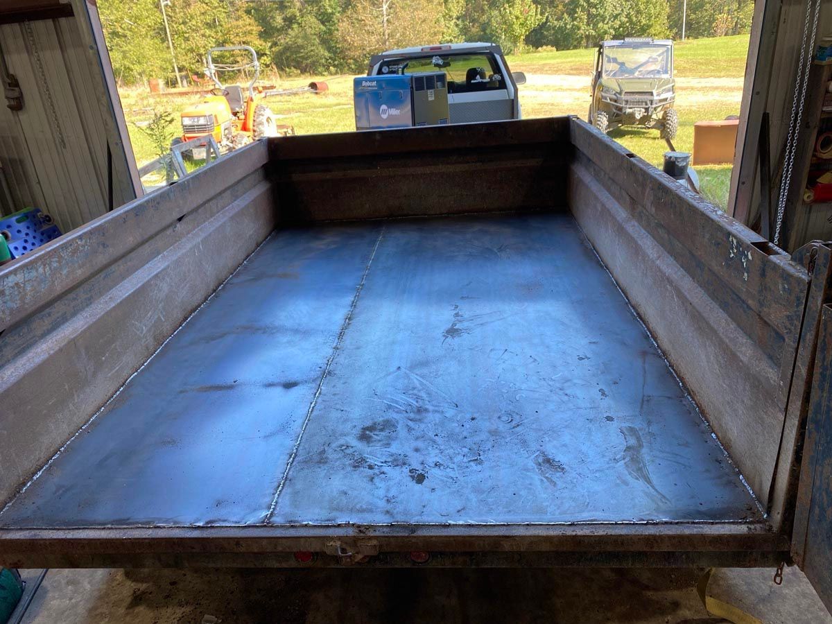 A metal-floored trailer bed with wooden side walls, viewed from behind in an outdoor workshop or yard setting.