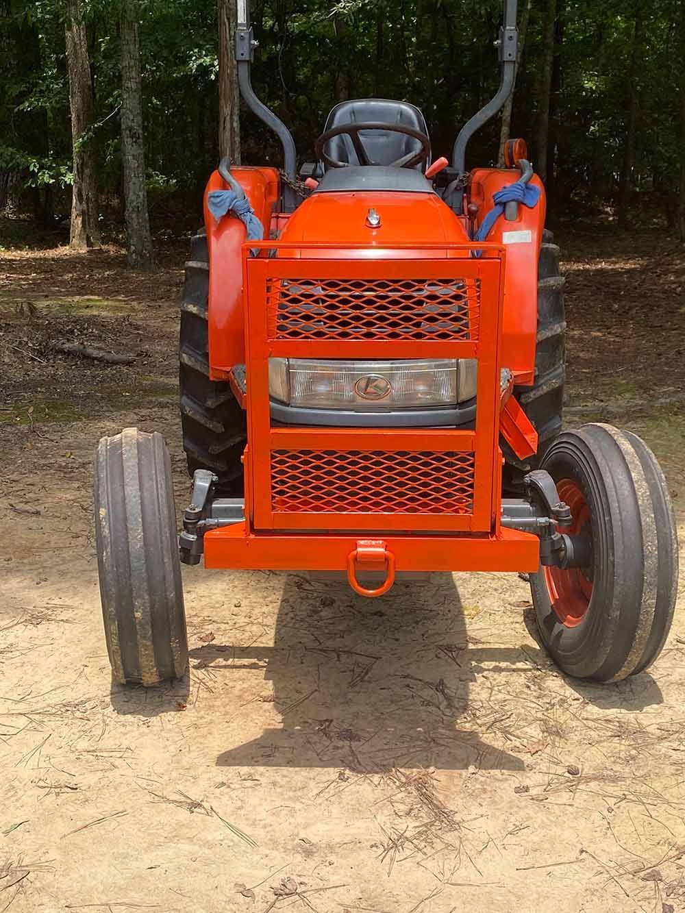 Orange tractor with a protective metal grille over the front hood and headlights, parked on a dirt path in the woods.
