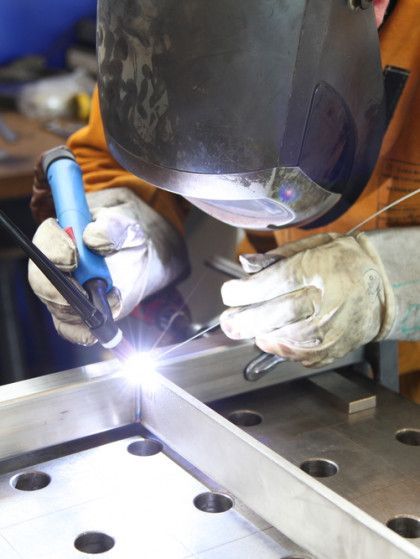 A welder in protective gear uses a TIG torch to join metal pieces on a perforated workstation table.
