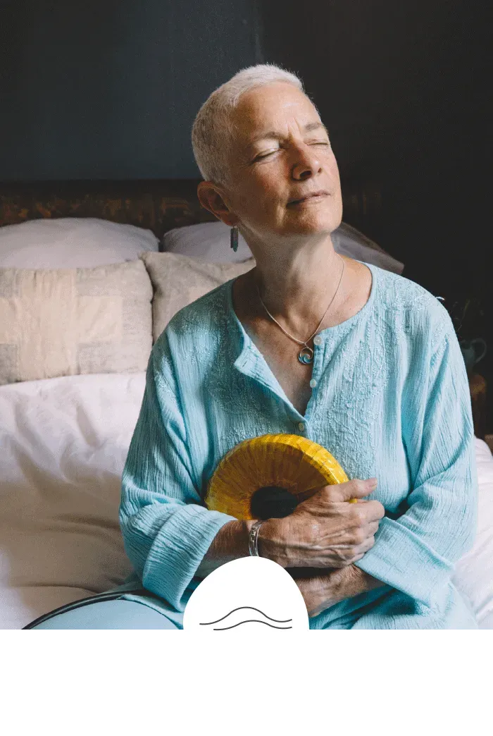 An elderly woman is sitting on a bed with her eyes closed and holding a yellow object.