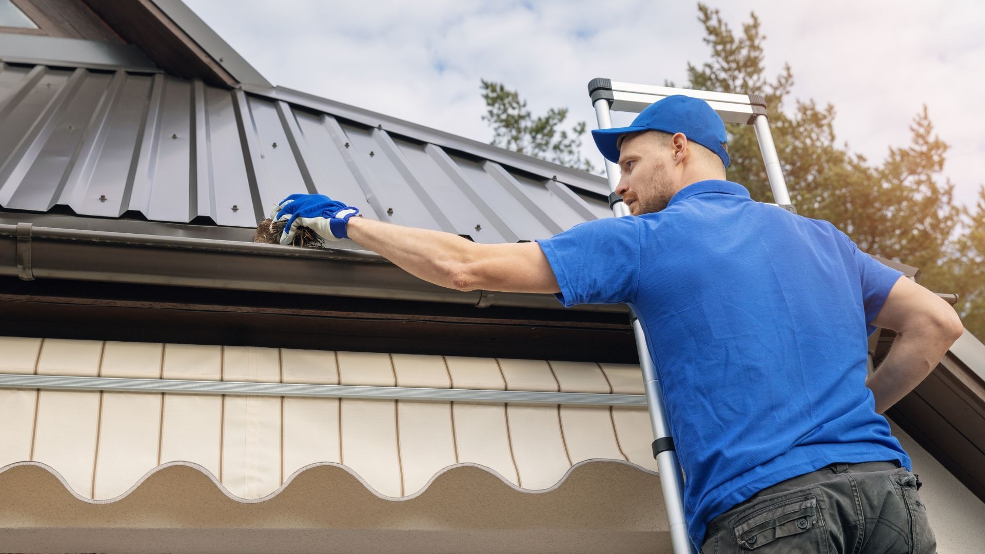 A man is standing on a ladder cleaning a gutter on the roof of a house.