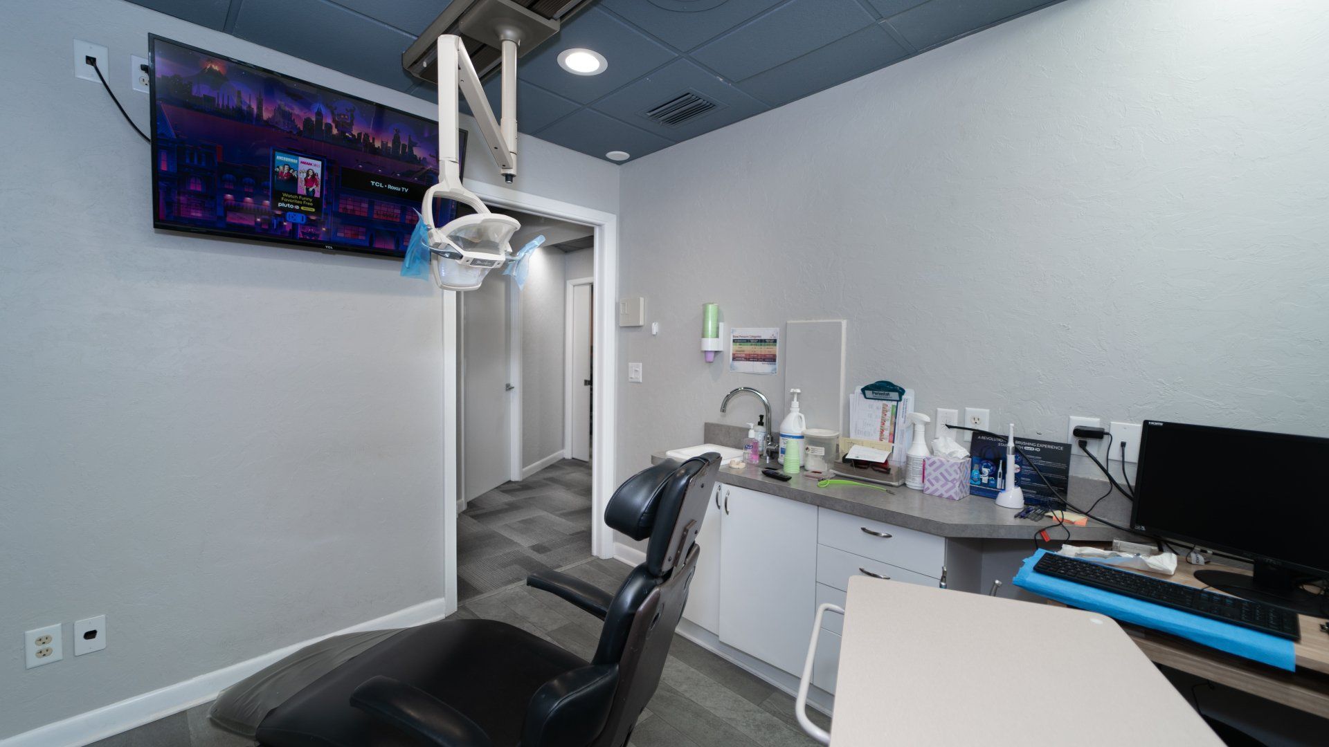 A dental office with a chair , desk , computer and television.