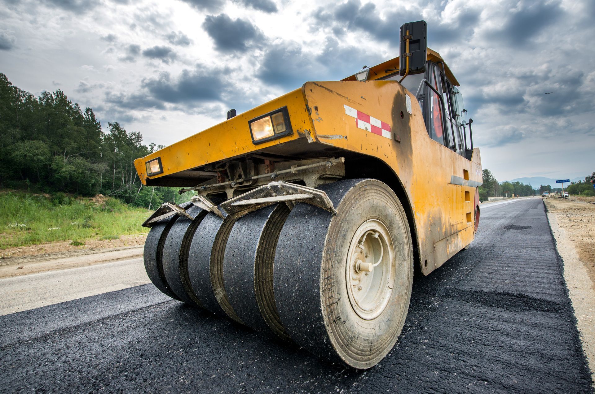 Yellow road roller compressing new asphalt on highway construction site under cloudy sky.