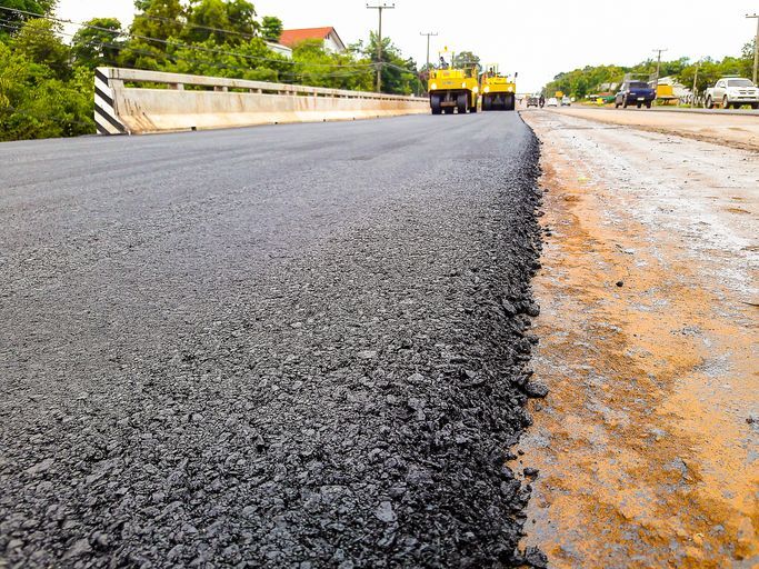 Freshly paved asphalt road near bridge with construction vehicles and greenery in background.