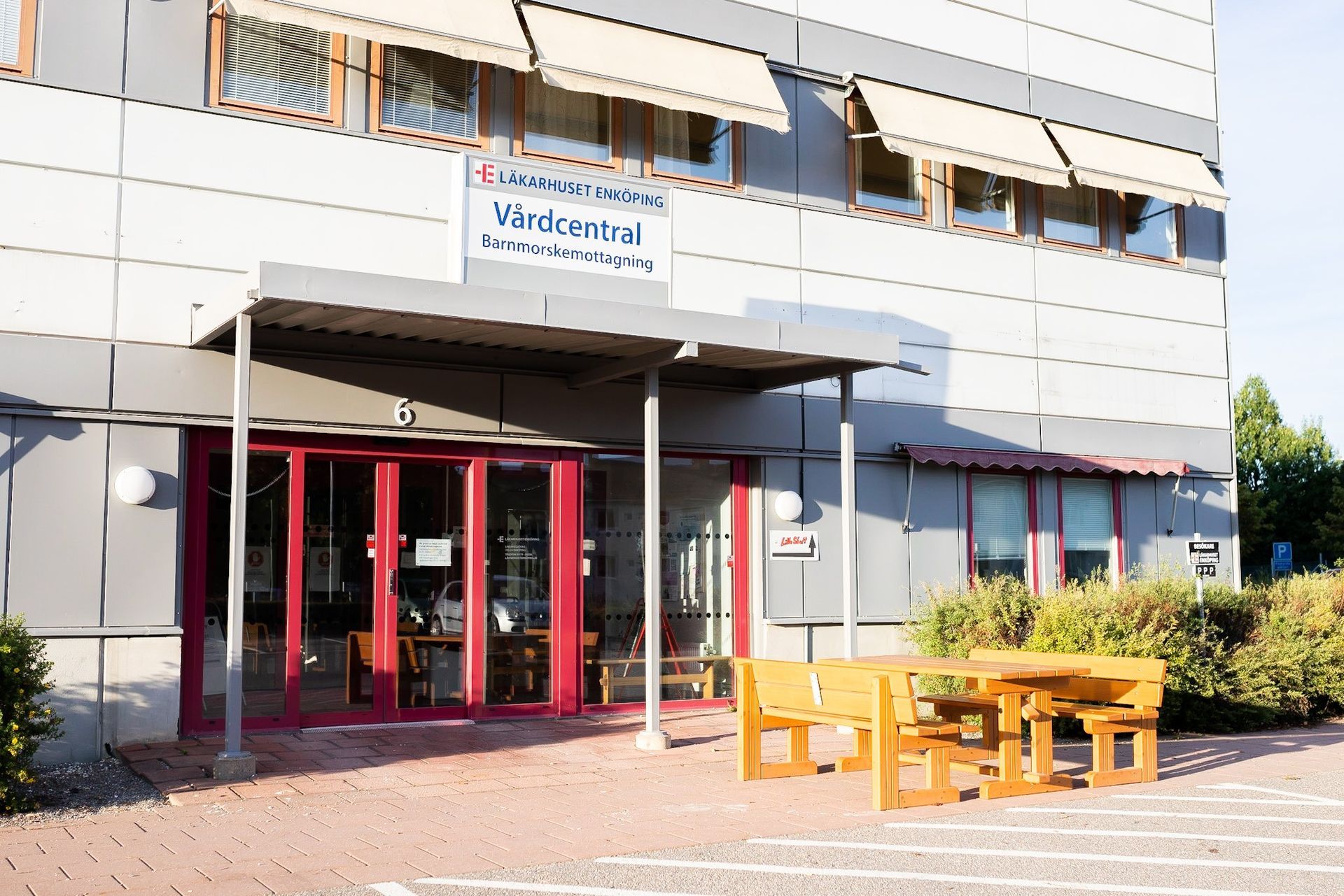 Ingång till en byggnad med röda dörrar och bänkar. Skylten säger 