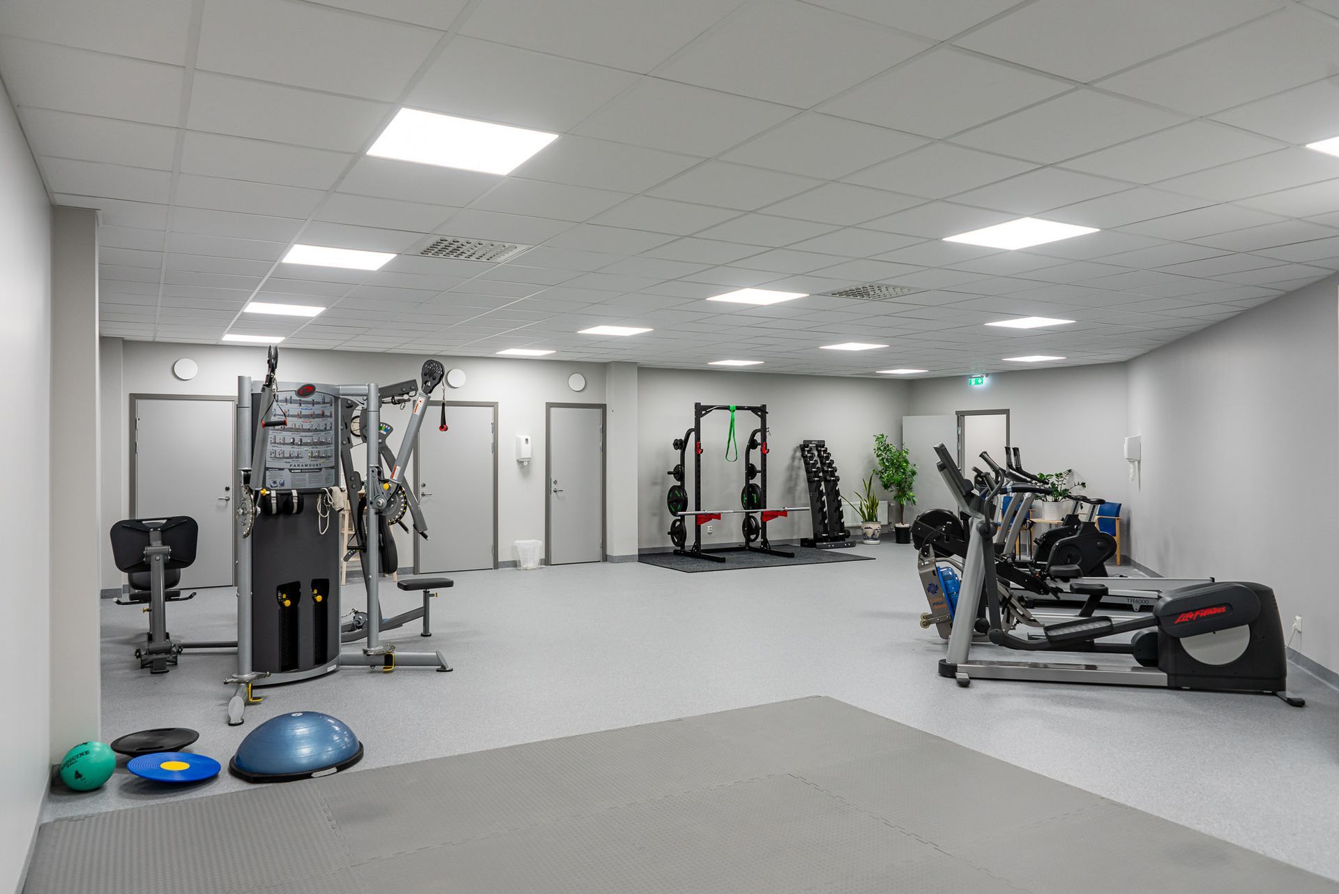 Gyminredning med träningsmaskiner, grått golv, vita väggar och tak samt flera dörrar.