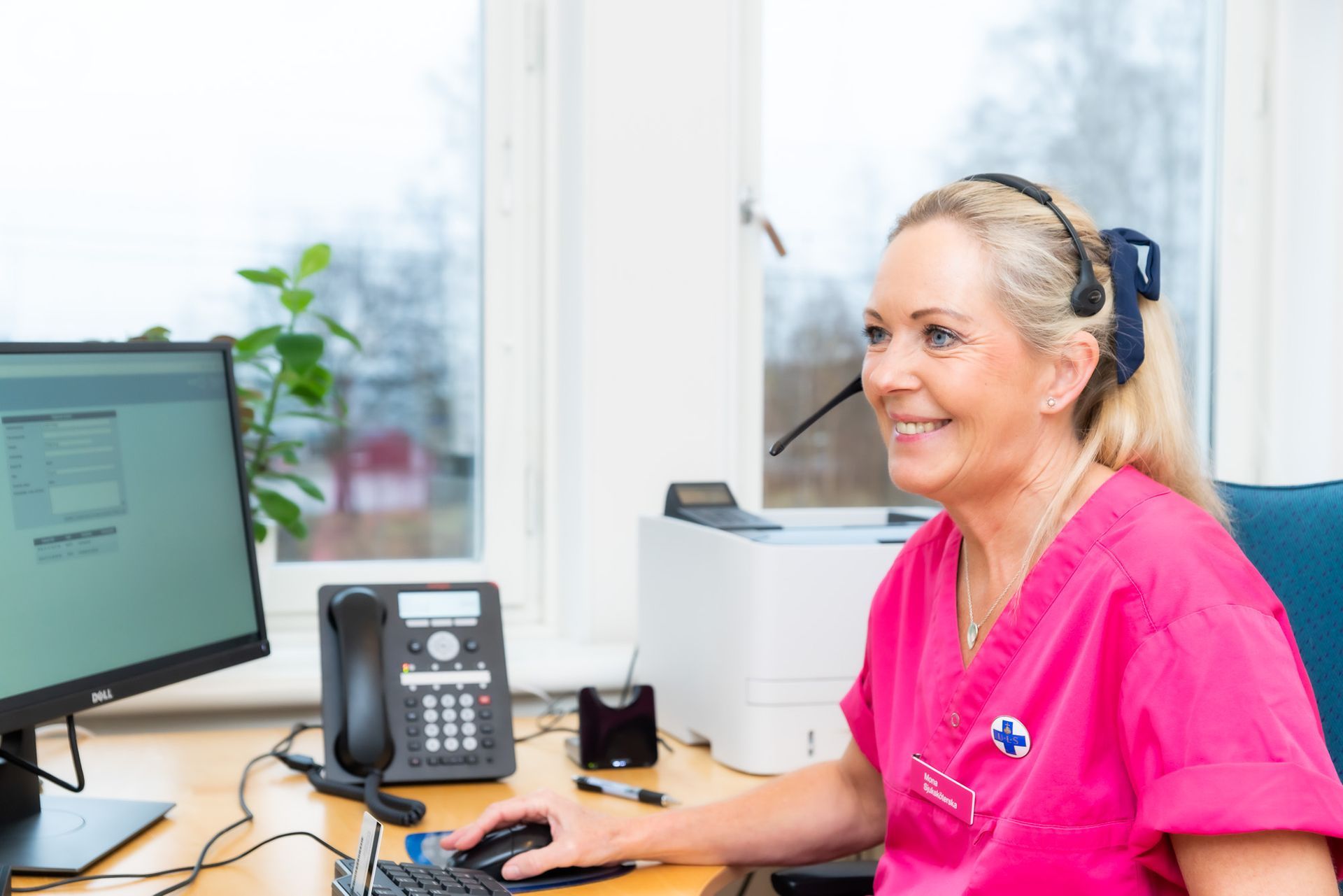 Kvinna med headset och rosa scrubs ler mot datorn i kontorsmiljö.