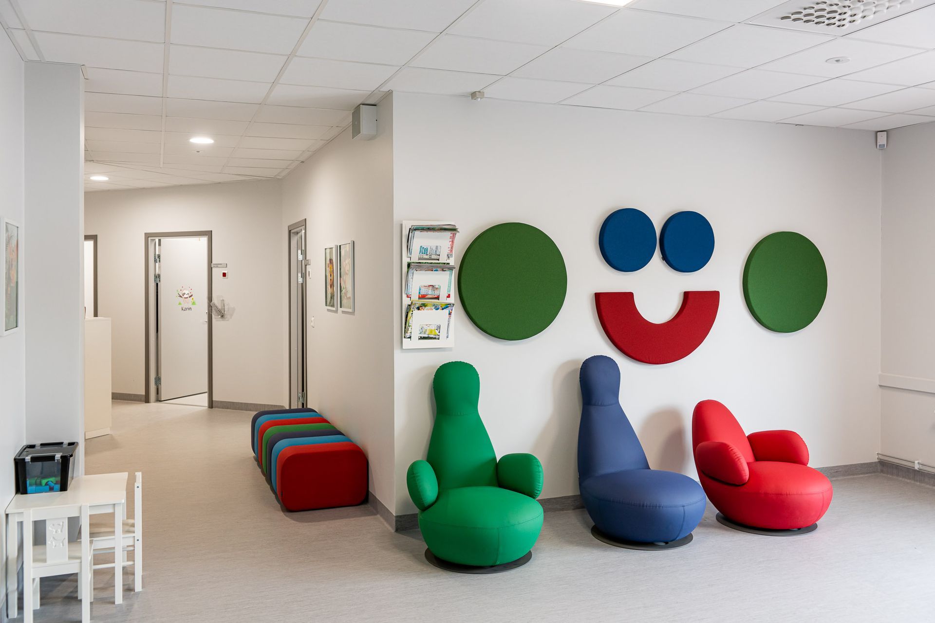 Väntrum med färgglada stolar och väggdekorationer; hallen leder till dörrar.