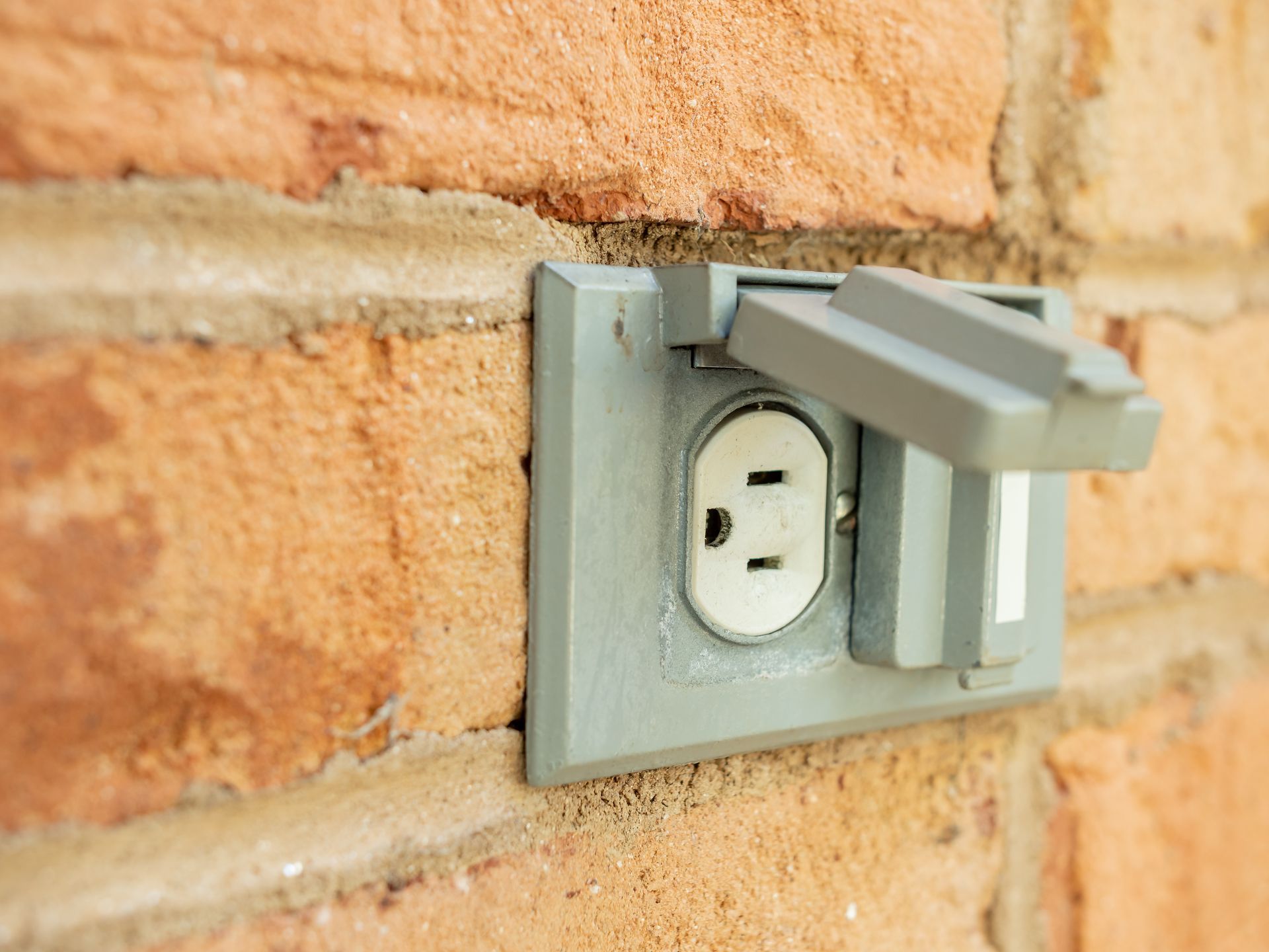 Outdoor electrical outlet with a gray weatherproof cover on a brick wall.