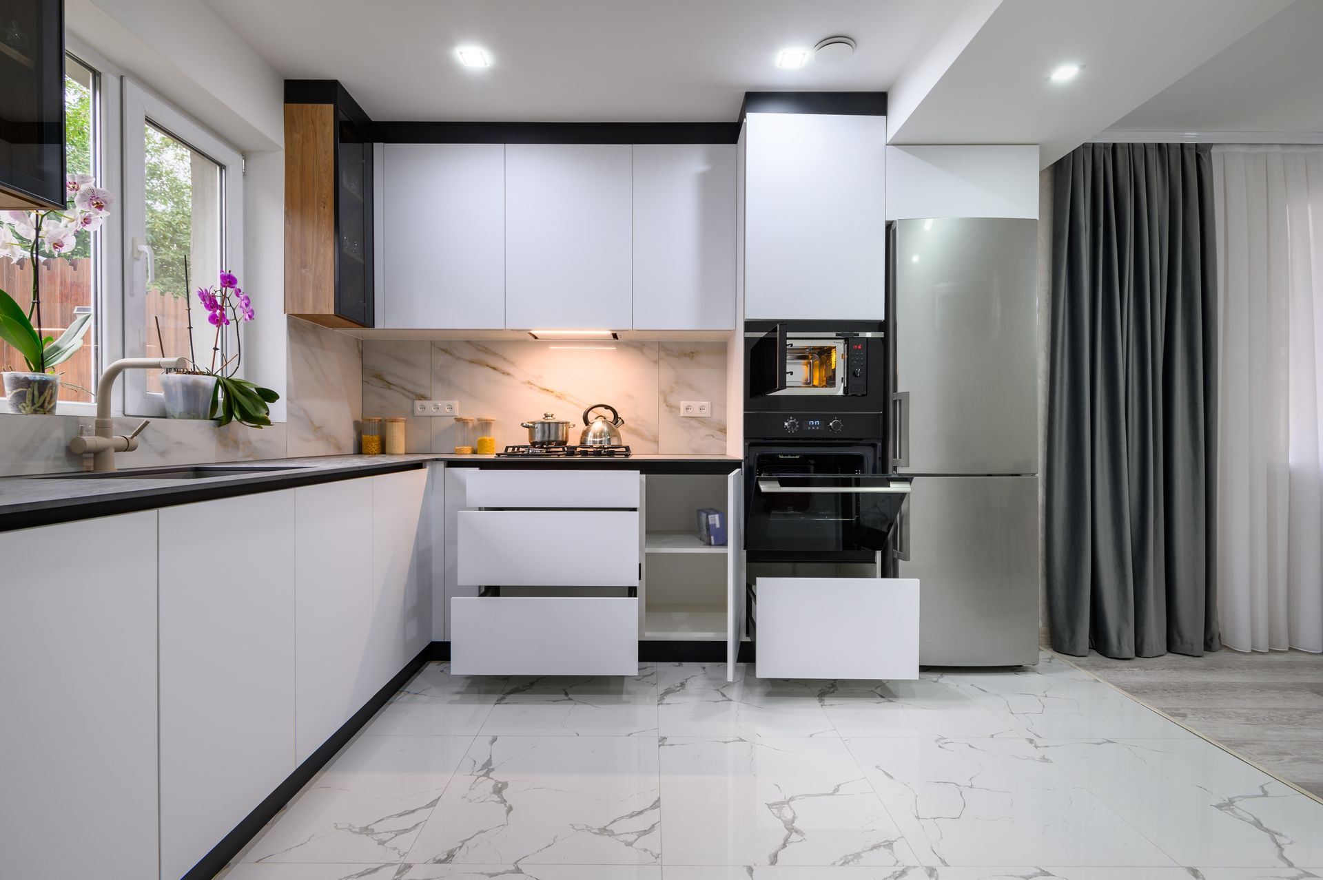 Modern white kitchen with marble countertops and backsplash, stainless steel appliances, and large window.