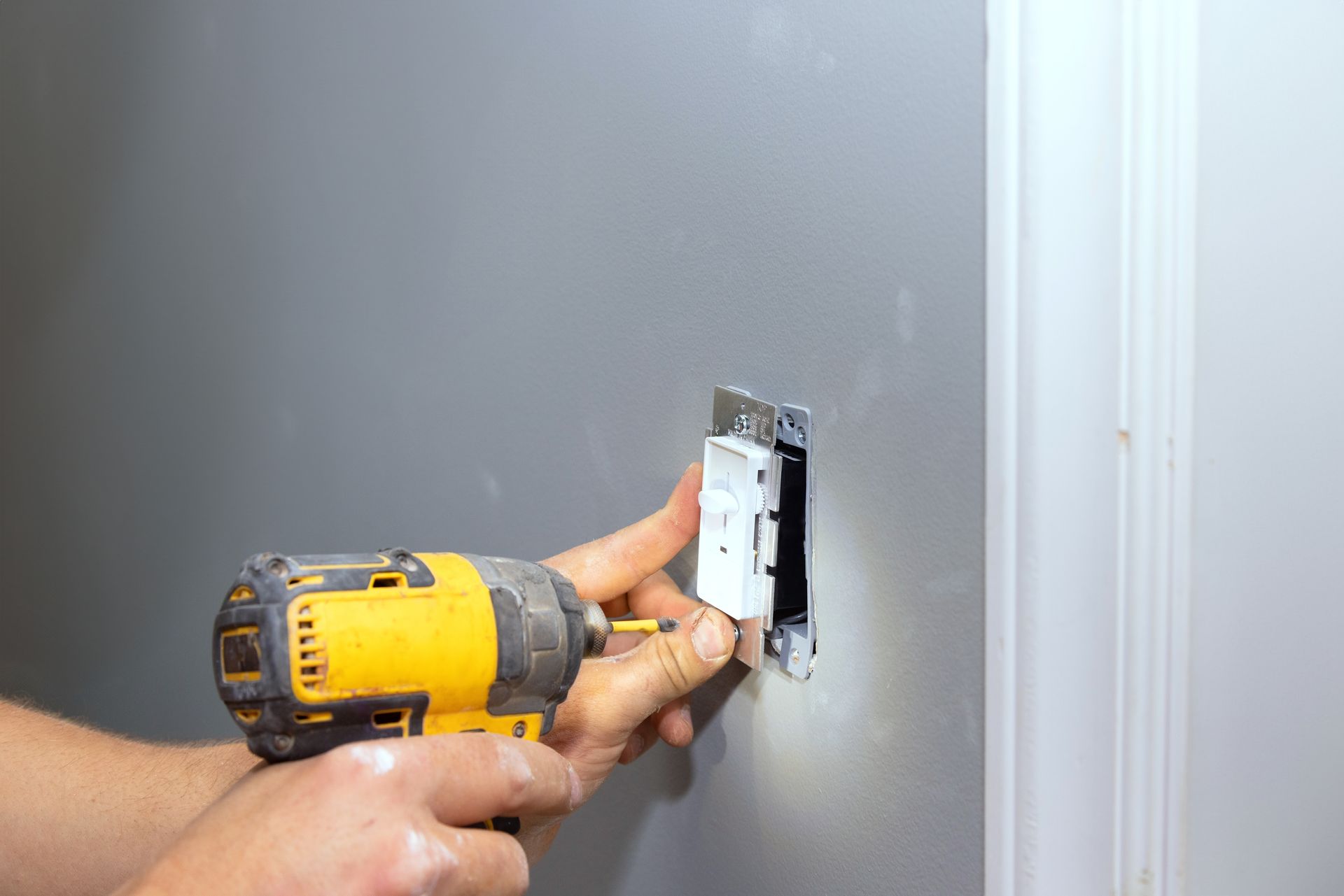 Person installing a light switch with a yellow drill on a gray wall.
