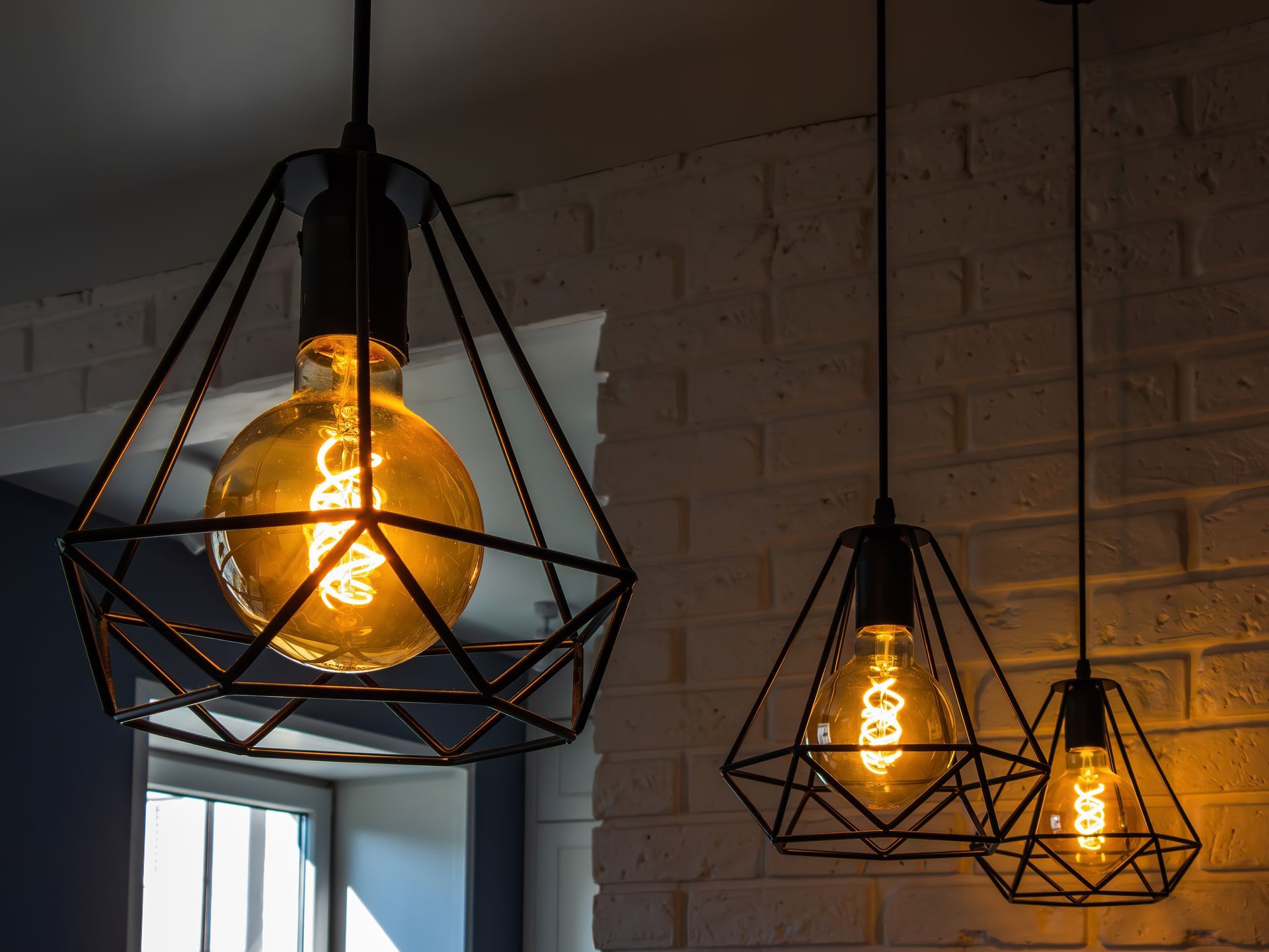 Three pendant lights with exposed, glowing bulbs encased in black geometric metal frames, near a white brick wall.