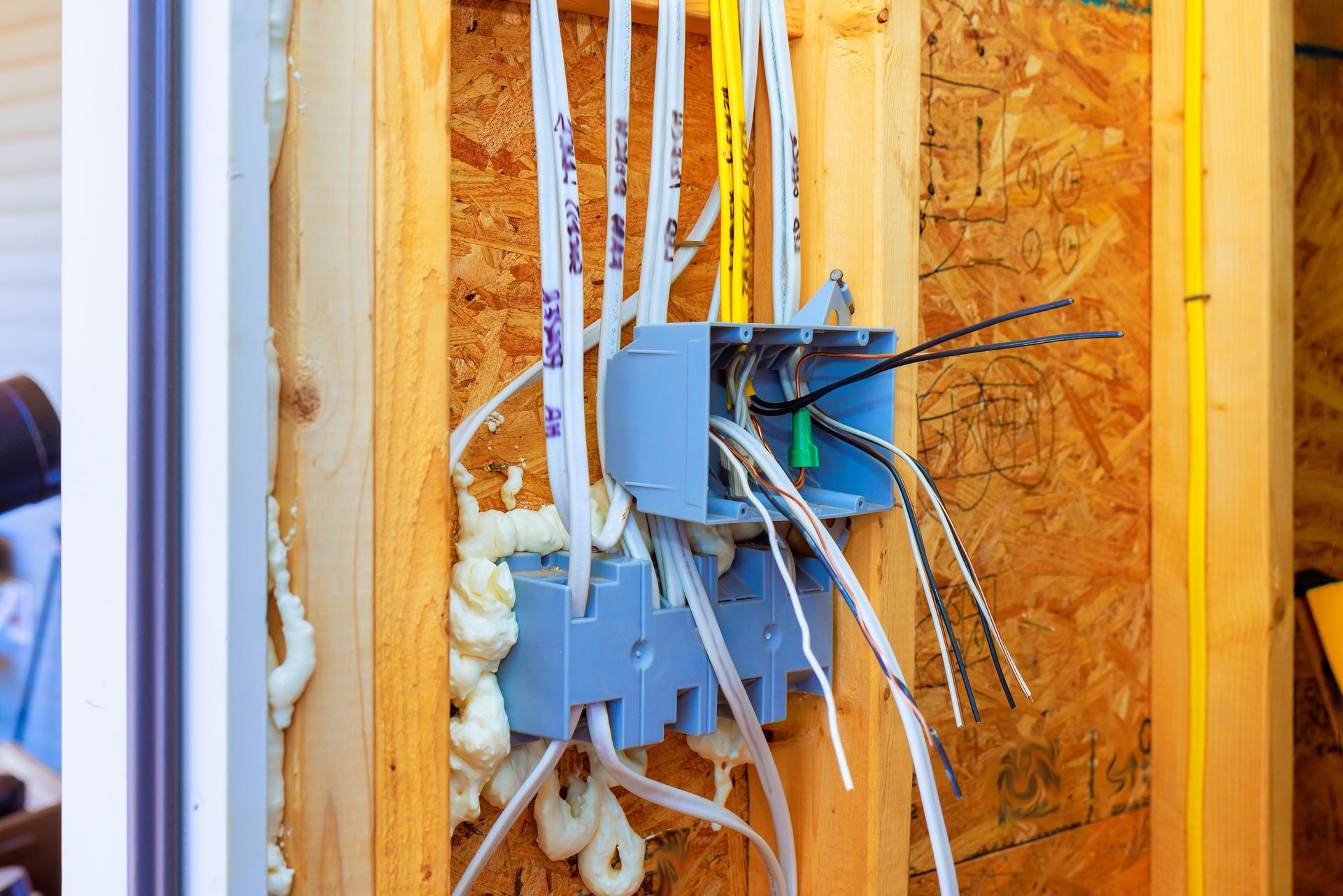 Blue electrical junction box with wires in wooden wall. Foam sealant surrounds the box and wires.
