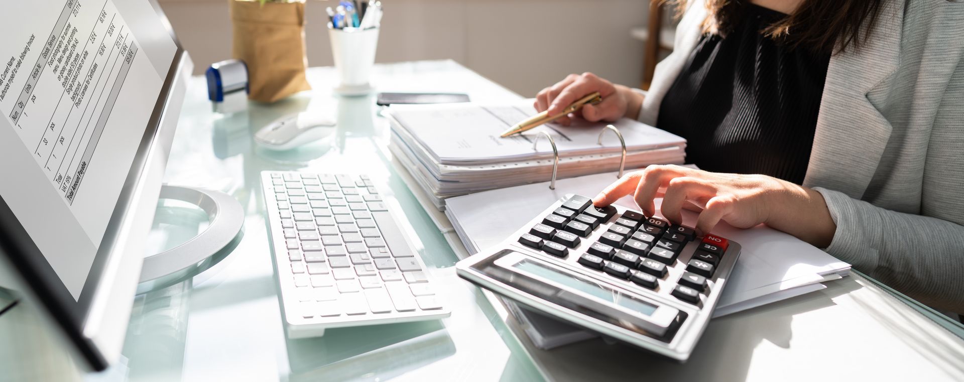 A woman is sitting at a desk using a calculator and a computer.
