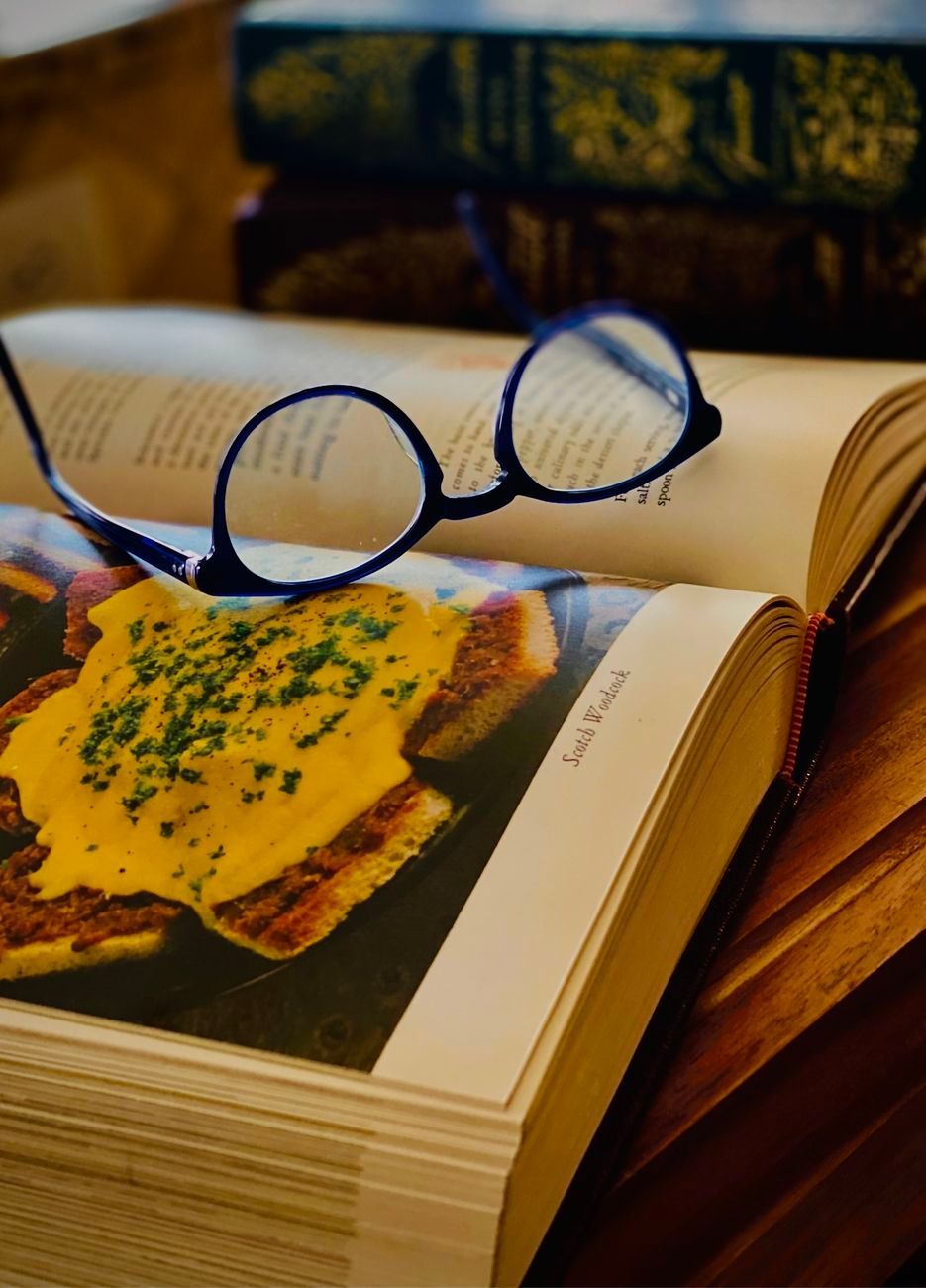 Eyeglasses on open cookbook with food photo; stack of books in background.