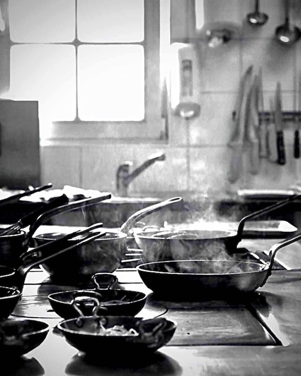 Kitchen scene with steaming pots on stove, sink, knives, and window in background.
