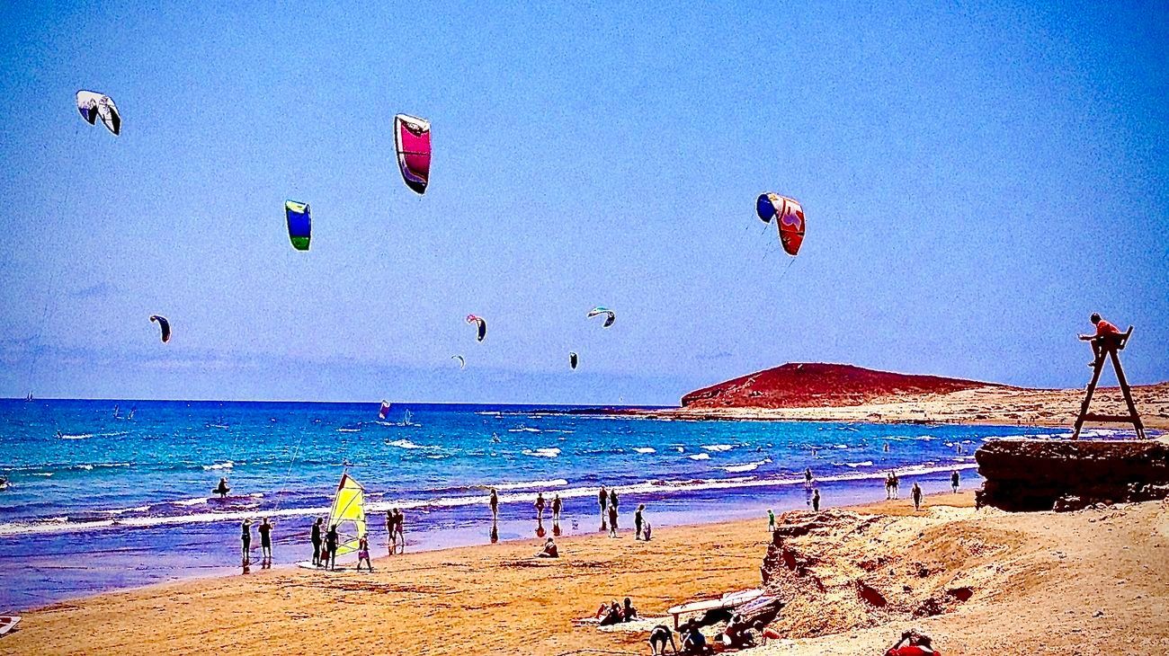 Kite surfers on a sunny beach with blue water and sky, a red hill in the distance.