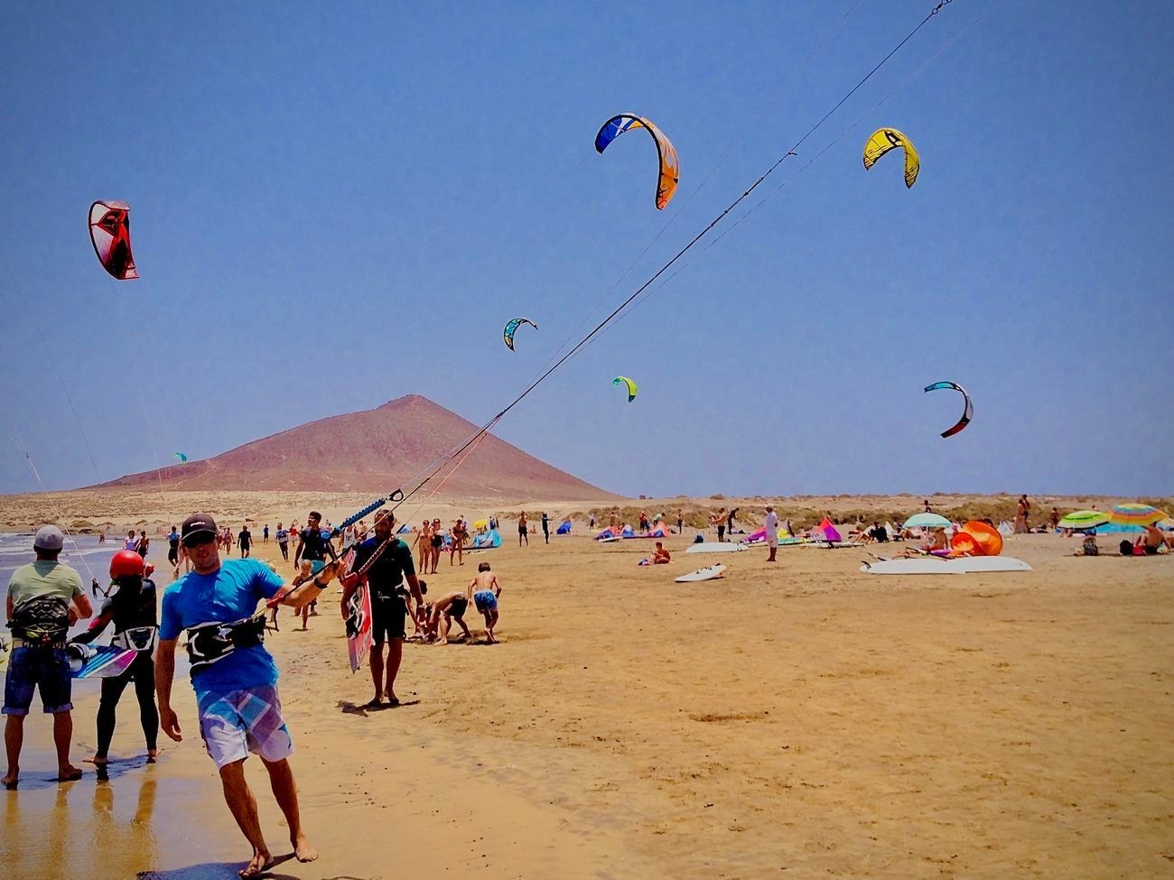Beach scene with kite surfers. People on sandy shore, kites in the sky, and a mountain in the background.