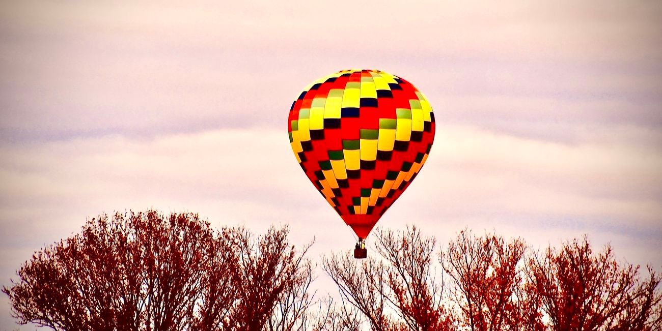 Colorful hot air balloon soaring above trees against a cloudy sky.