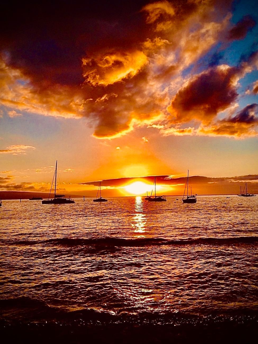 Sunset over ocean with boats silhouetted on water. Fiery orange and yellow clouds reflect on water's surface.