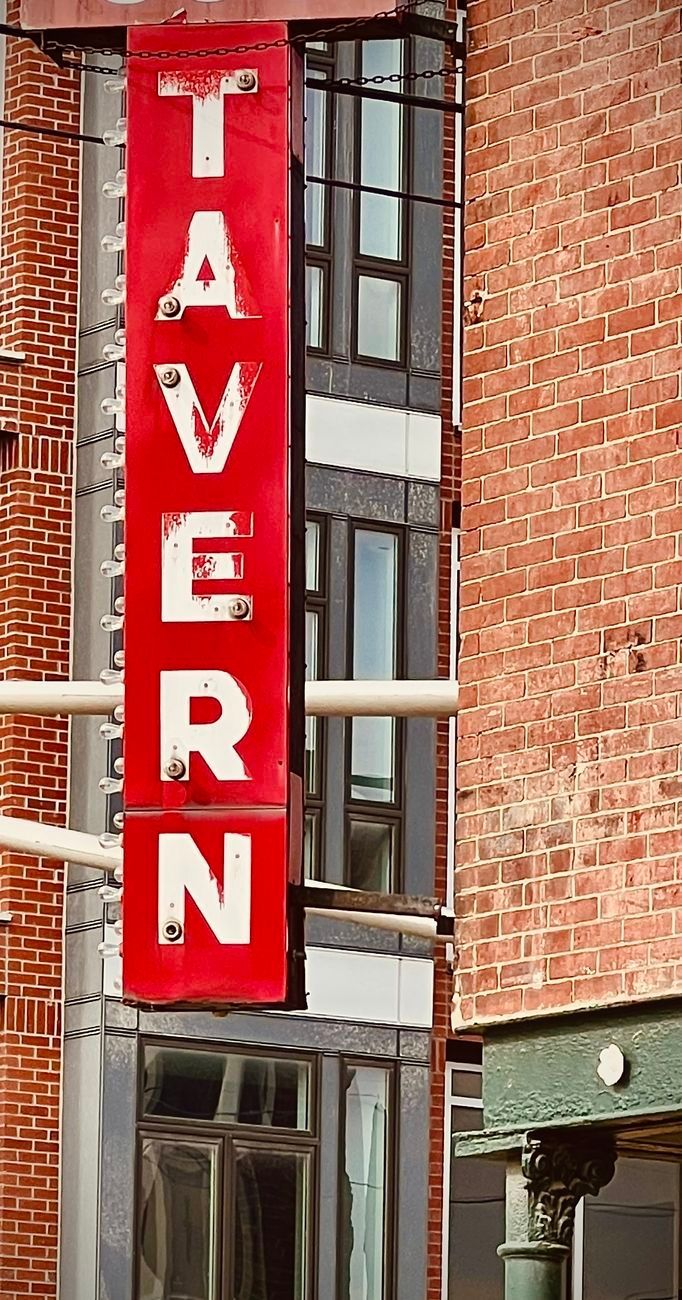 Red tavern sign with white letters on a brick building facade.