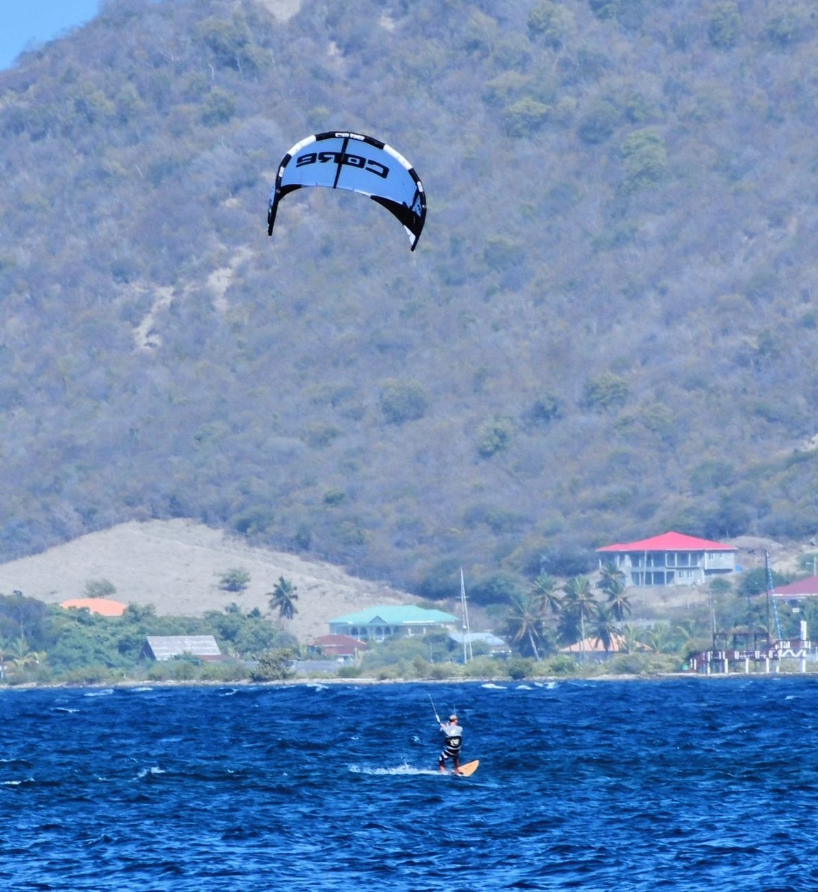 Kitesurfer on blue water with a mountain in the background. The kite is light blue and black.