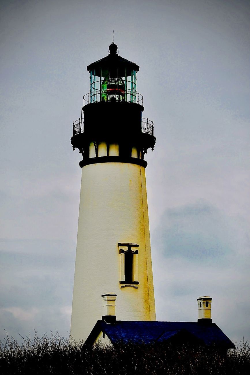 White lighthouse with black top, green light, and blue roof against a cloudy sky.