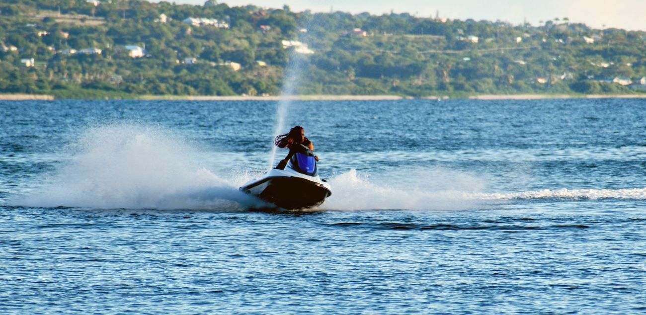 A person riding a jet ski on blue water, creating white spray, with a green, hilly background.