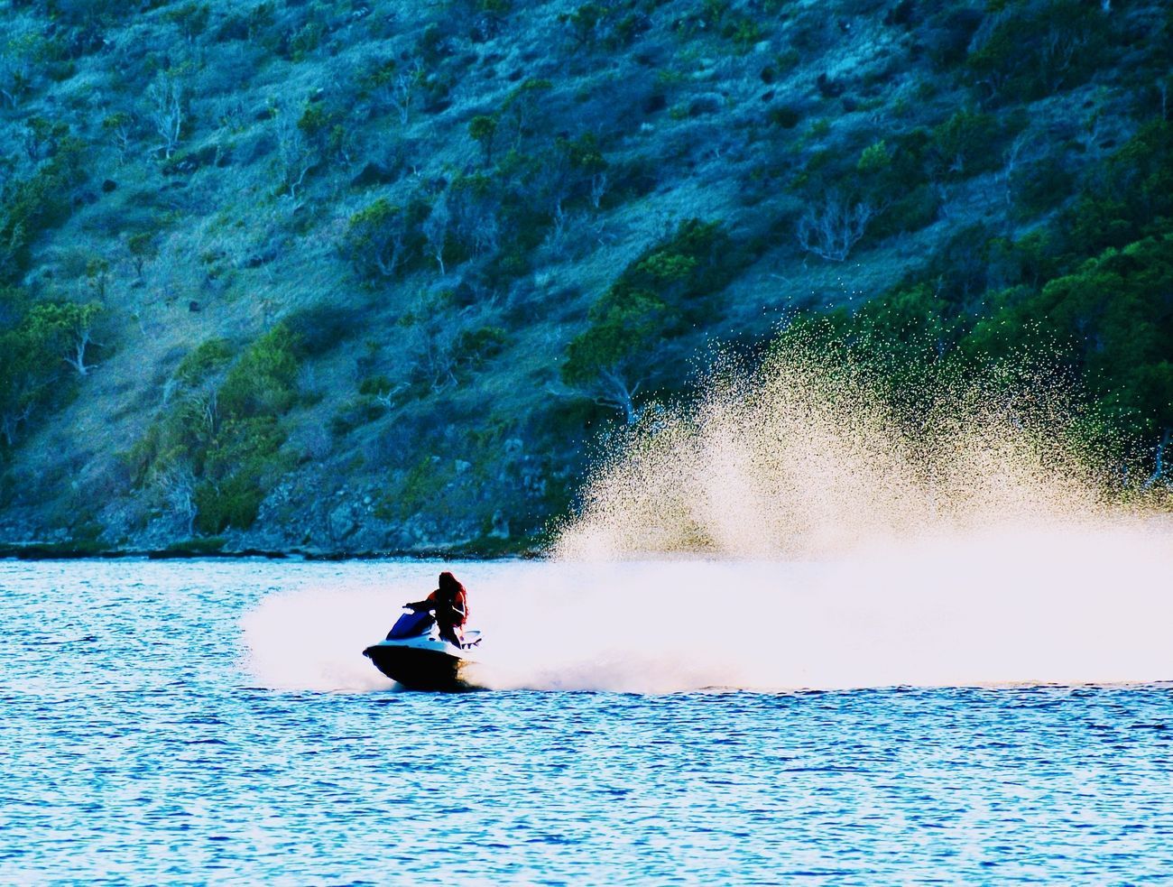 Jet skier on water, creating a large spray. Mountainous, tree-lined background, blue water.