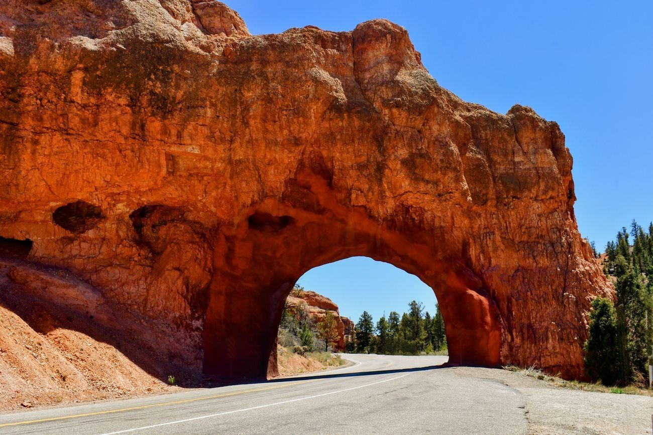 Road passing through a natural red rock arch under a bright blue sky.