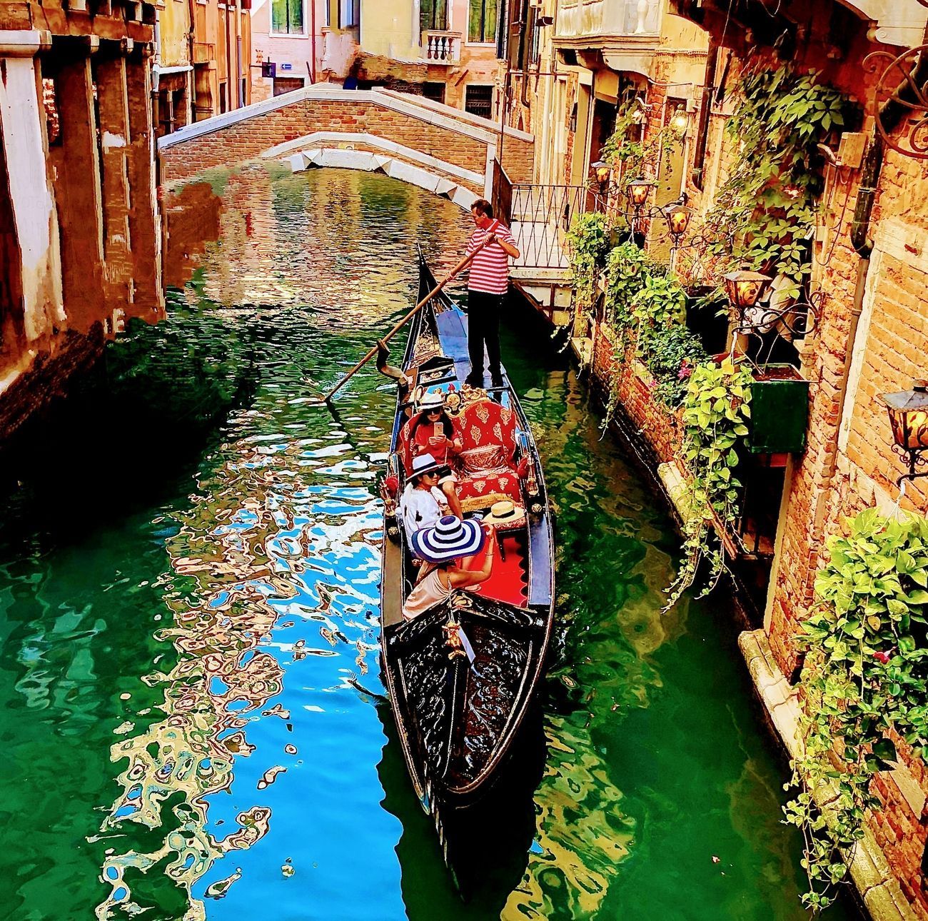 Gondola carrying tourists travels along a Venetian canal, under a small stone bridge, lush greenery on the walls.