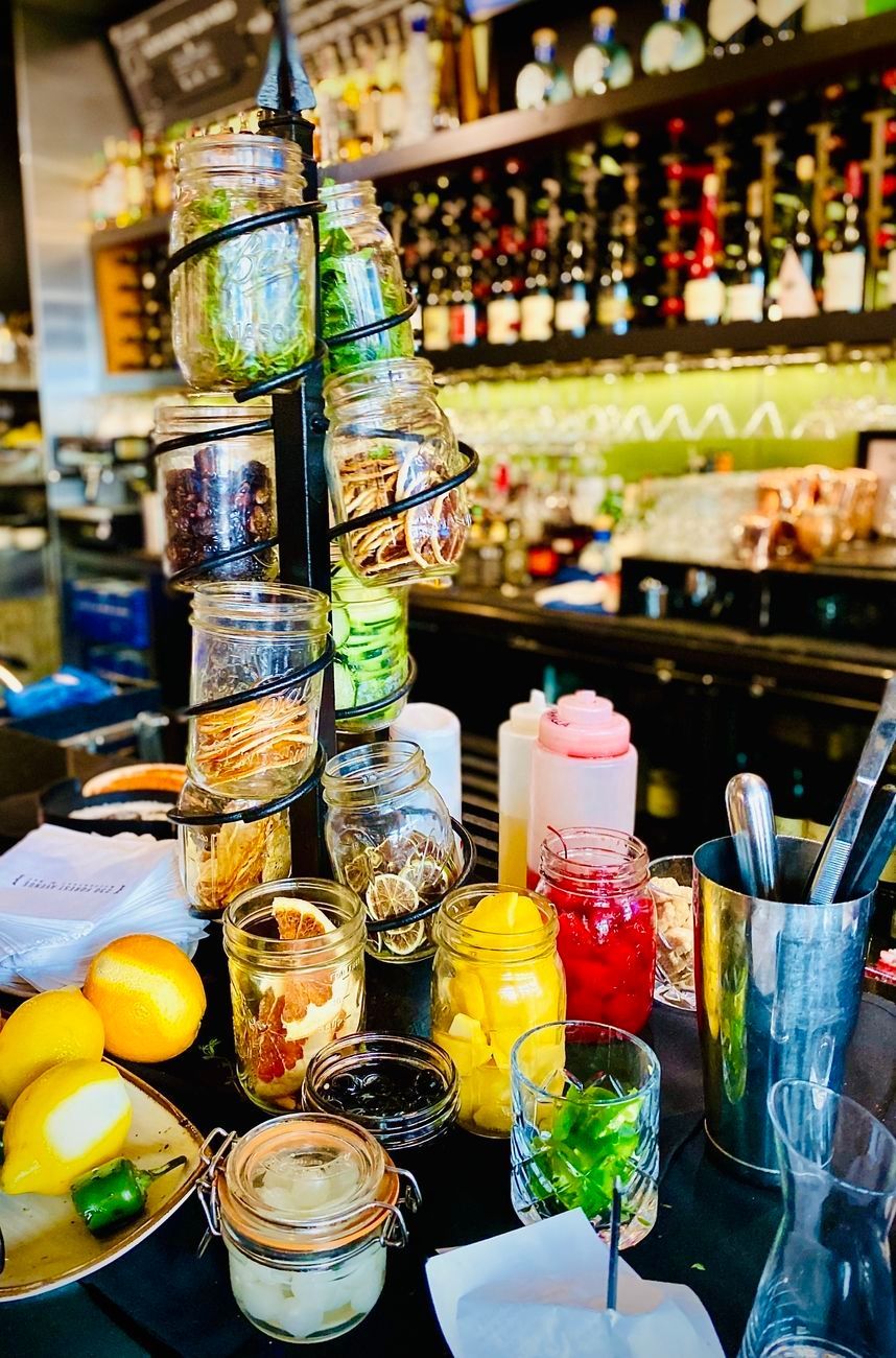 Bar setup with jars of garnishes and colorful syrups. A selection of citrus fruits are visible.