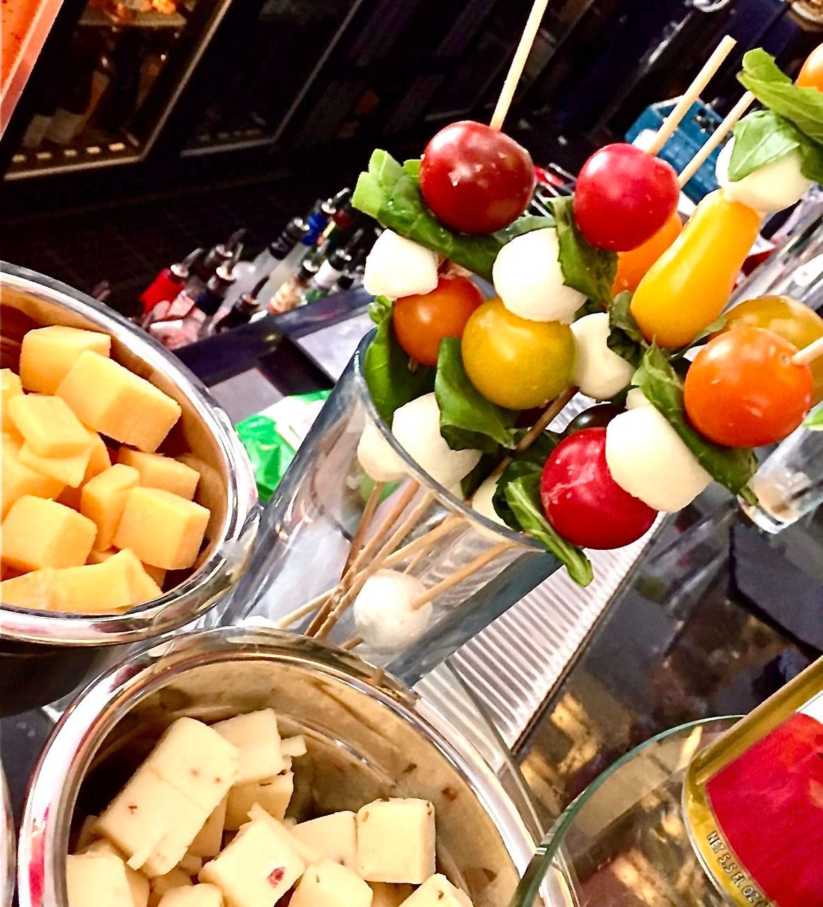 Skewers of cherry tomatoes, mozzarella, and basil in a glass, with cheese cubes in silver bowls.