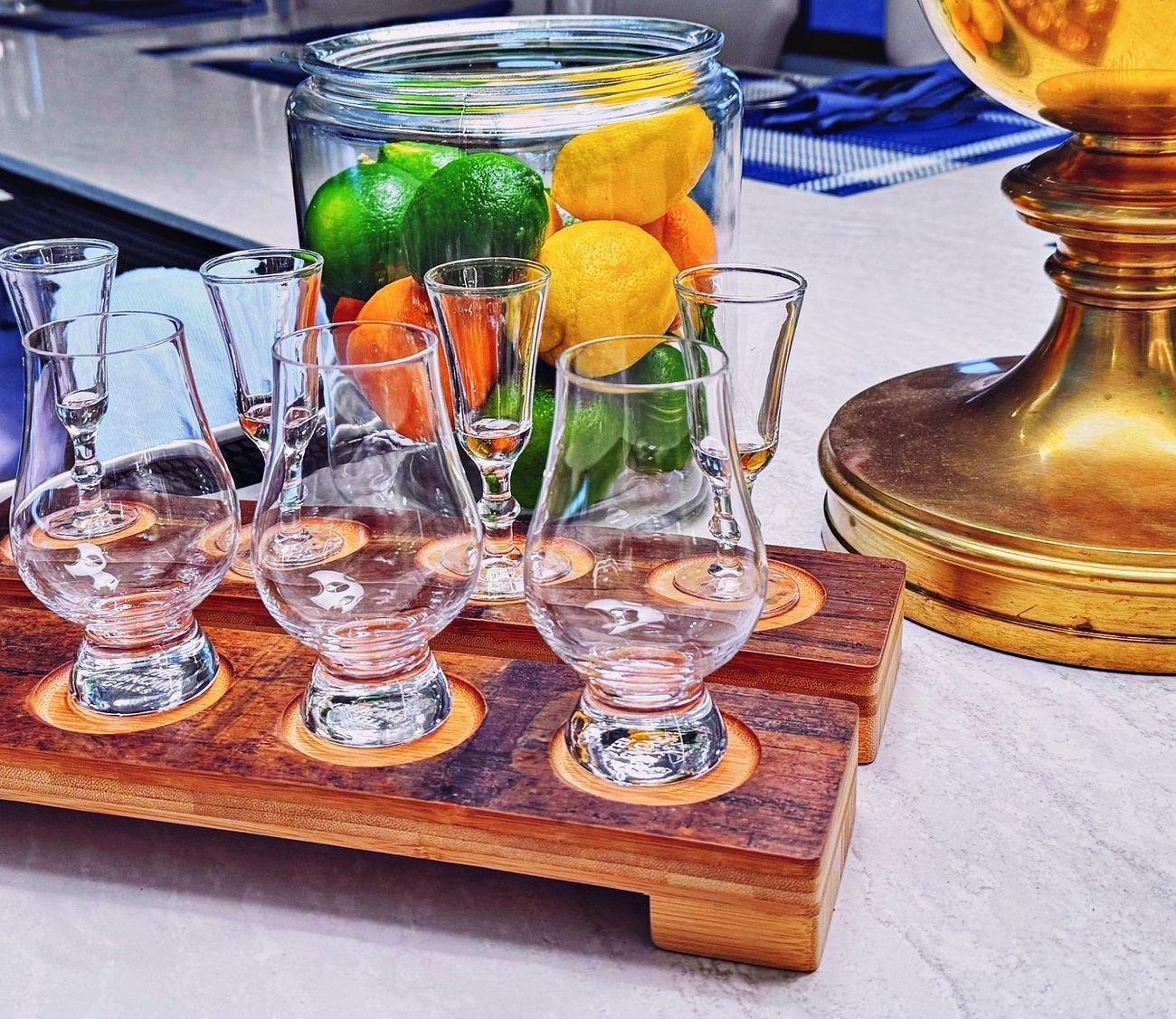 Whiskey tasting flight with clear glasses on wooden tray, citrus fruits in jar, gold lamp base.
