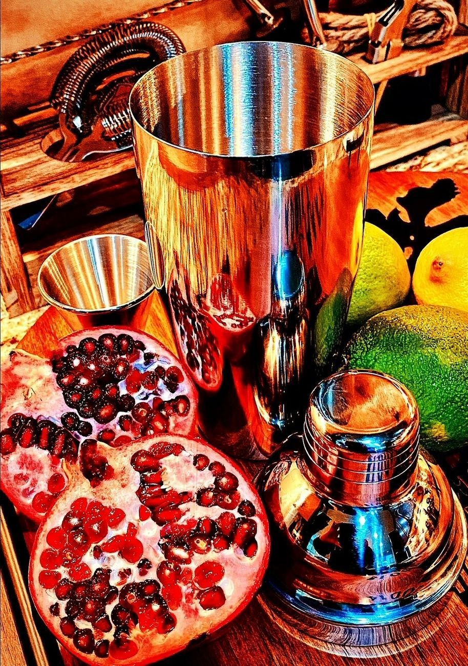 Cocktail shaker with half-cut pomegranate, limes, and lemons; close-up. Shiny metal with bright colors.