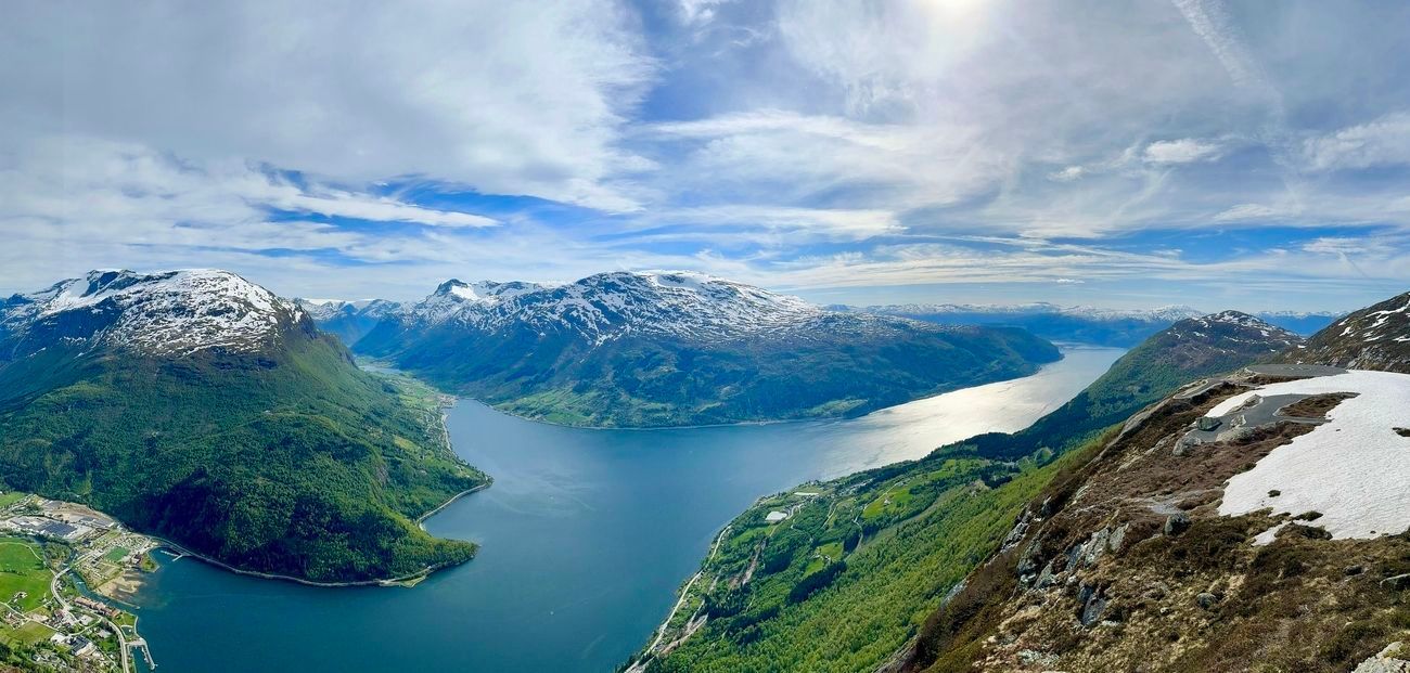 Panoramic view of a fjord with mountains covered in snow and lush green vegetation. Blue water and sky.