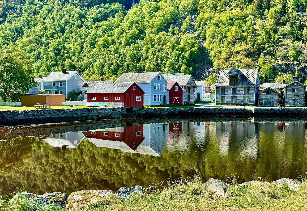 Colorful buildings reflect in calm water; green trees cover hills in Norway.